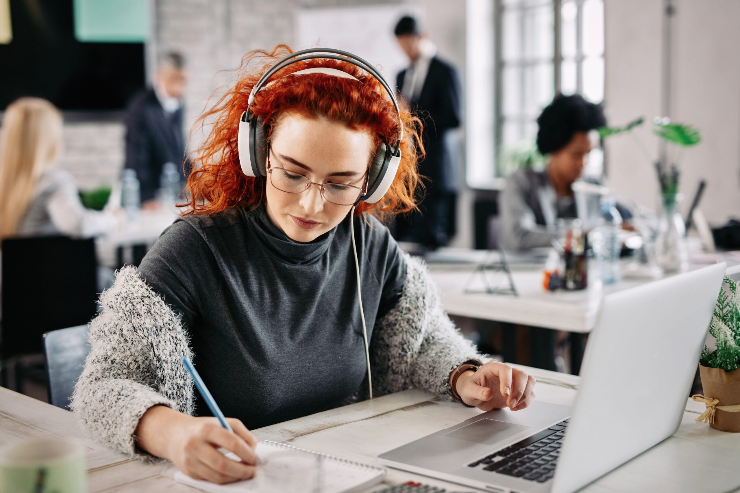Redhead businesswoman using laptop and writing notes in her notepad while listening music on headphones at work.